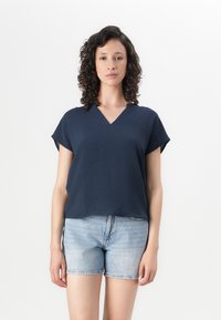 Marineblaue, kurzärmlige V-Ausschnitt-Bluse mit strukturierter Oberfläche, kombiniert mit hellen Denim-Shorts. Lockere Passform und lässiges Design.