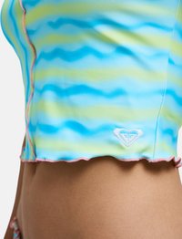 Κοντινό πλάνο του κορμού μιας γυναίκας που φοράει ένα cropped top με κυματιστό μπλε και πράσινο μοτίβο, με ροζ περίγραμμα και μικρό λευκό κεντητό λογότυπο κοντά στο τελείωμα.