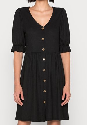 Woman wearing a black button-down dress with puff sleeves and ruffled cuffs, standing against a plain background.