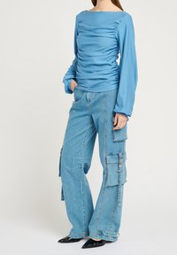 Blouse bleue à manches longues avec des détails froncés, associée à un pantalon cargo en denim bleu à jambes larges, doté de multiples poches et d'une taille haute.
