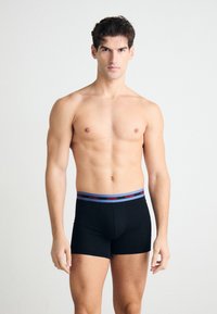 Boxers noirs avec une ceinture bleu et rouge portant l'inscription "HUGO". Tissu lisse, design ajusté, sans motifs ni accents visibles. Le modèle se tient droit.