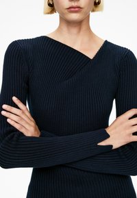 Frau mit kurzen blonden Haaren, die einen navyblauen RIPPSWIE Wolle trägt, mit verschränkten Armen und einem Schlitzdetail am Daumen des Ärmels.