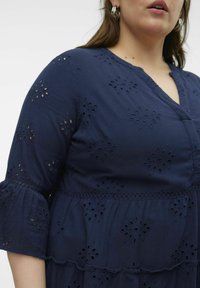 navyblaues Kleid aus strukturiertem Stoff mit bestickten floralen Ausschnitten, einem schulterfreien Ärmel und einem gestuften Design.
