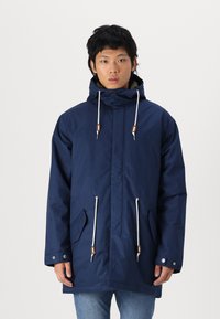 Marineblauwe waterdichte parka met een capuchon, voorzien van witte trekkoorden met bruine accenten, voorzakken en een rechte snit.