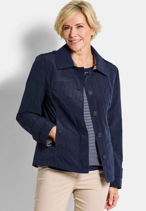 Femme blonde souriante, portant une veste marine boutonnée par-dessus une chemise rayée et un pantalon beige, avec une main dans la poche de la veste.