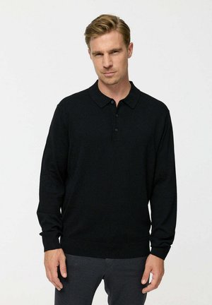 Mann trägt ein schwarzes Langarm-Polohemd und dunkelgraue Hosen, steht vor einem einfarbigen weißen Hintergrund.