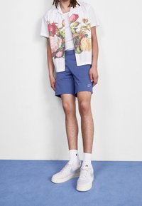 Vit kortärmad skjorta med blommönster, i kombination med blå shorts och vita Nike-sneakers. Blå bakgrund, minimalistisk design.
