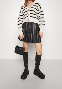 Randig gräddfärgad och svart stickad cardigan med stora knappar, i kombination med en veckad svart faux läderkjol, höga svarta plattformstövlar, och en liten väska.