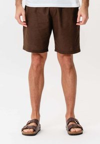 Mand iført brune shorts og mørkebrune sandaler står mod en hvid baggrund med hænderne afslappet ved siden af.