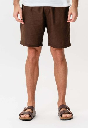 Mand iført brune shorts og mørkebrune sandaler står mod en hvid baggrund med hænderne afslappet ved siden af.