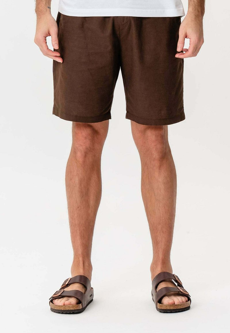 Mand iført brune shorts og mørkebrune sandaler står mod en hvid baggrund med hænderne afslappet ved siden af.