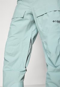 Pantaloni impermeabili de culoare teal deschis, cu ventilație laterală cu fermoar și buzunare mari, realizați dintr-un material durabil, cu o ajustare confortabilă și branding minim.
