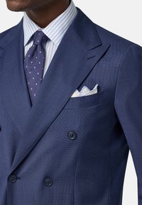 Completo blu navy doppiopetto in tessuto testurizzato, con rever a punta, bottoni scuri e una camicia a righe azzurro chiaro sotto. Fazzoletto da taschino visibile.