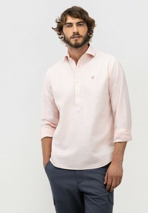 Hombre con barba y cabello ondulado que lleva una camisa de manga larga color rosa claro abotonada con las mangas remangadas y pantalones gris oscuro.