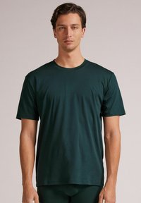 Ciemnozielona koszulka z krótkim rękawem, uszyta z gładkiego materiału, z okrągłym dekoltem i luźnym krojem, noszona na dopasowanych spodniach sportowych.