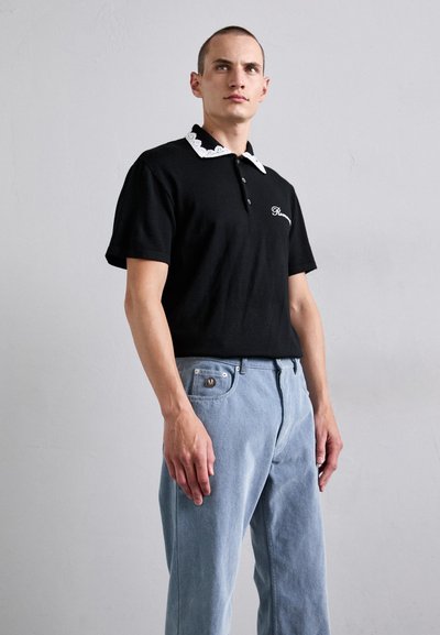 Bărbat îmbrăcat cu un polo negru cu mânecă scurtă și guler din dantelă albă, purtând blugi deschiși la culoare, stând în fața unui zid gri simplu.