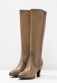 Botas de cuero marrón hasta la rodilla con un acabado suave y un acento textil acanalado en la parte posterior; presenta un tacón afilado y puntera redonda.