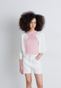 Cárdigan blanco sobre una camiseta rosa de manga corta con un pequeño logo, combinado con pantalones cortos de denim blancos. Tejido suave y corte relajado.