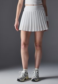 Jupe plissée blanche avec une taille haute, fabriquée en tissu lisse. Associée à des chaussures de sport grises et des chaussettes blanches à bord côtelé.