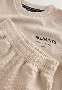 Beež värvusega AllSaints sametise waffle-keerdumisega mugavusriiete komplekt, millel on elastne vööümbermõõduga püksid ja ümar kaelusega top, millel must logo ja jaapani tekst.