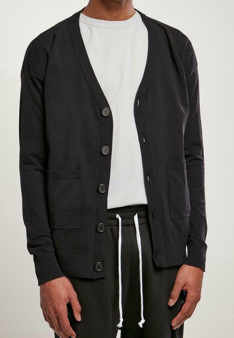 Svart långärmad cardigan med knappar, med ribbade ärmslut och V-ringning. Inkluderar sidofickor och är lager på en vit skjorta.