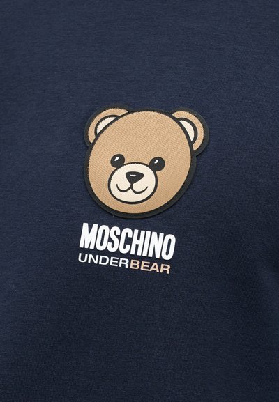 Μπαλωμάκι με το πρόσωπο ενός χαμογελαστού αρκούδου πάνω από το κείμενο "MOSCHINO UNDERBEAR" σε σκουρόχρωμο ναυτικό ύφασμα, πιθανώς μέρος ρούχου.