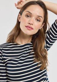 Maglietta a maniche lunghe a righe blu navy e bianche con scollo rotondo, design aderente e texture morbida. La modella posa con la mano dietro la testa.