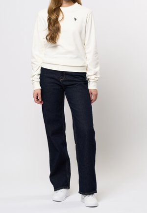 Krämfärgad sweatshirt med rund hals och långa ärmar, kombinerad med mörkblå rakt skurna jeans och vita sneakers. Enkel broderad logotypdetalj.