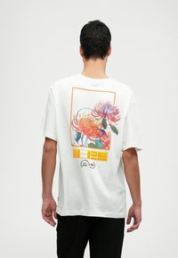 Camiseta de algodón blanca con un gráfico floral colorido en la parte posterior que presenta acentos en naranja y azul, marco rectangular y detalles del logotipo.