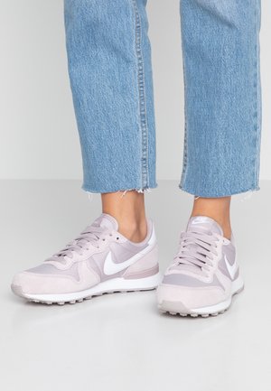 Person iført lyse lilla og hvide Nike-sneakers med snørebånd, kombineret med korte lyseblå denimjeans på en grå overflade.