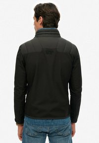 Homme portant une veste Superdry noire et un jean bleu, vu de dos, debout contre un fond blanc uni.