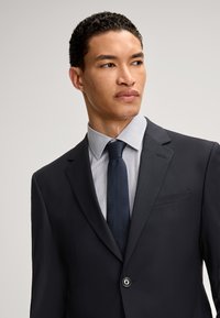 Dunkelblaues maßgeschneidertes Sakko mit einem einzigen Knopf, getragen über einem hellgrauen karierten Hemd und einer navyblauen Krawatte; glatte Textur und elegantes Design.