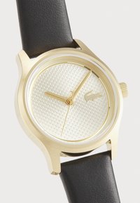 El reloj de tono dorado presenta un dial plateado texturizado, una correa de cuero negro y un diseño minimalista con marcadores de horas sencillos y detalle del logotipo.