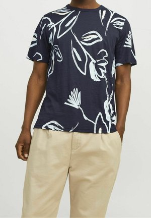 Homme portant un t-shirt bleu marine à manches courtes avec un motif abstrait de feuilles blanches et un pantalon beige, debout les mains dans les poches.