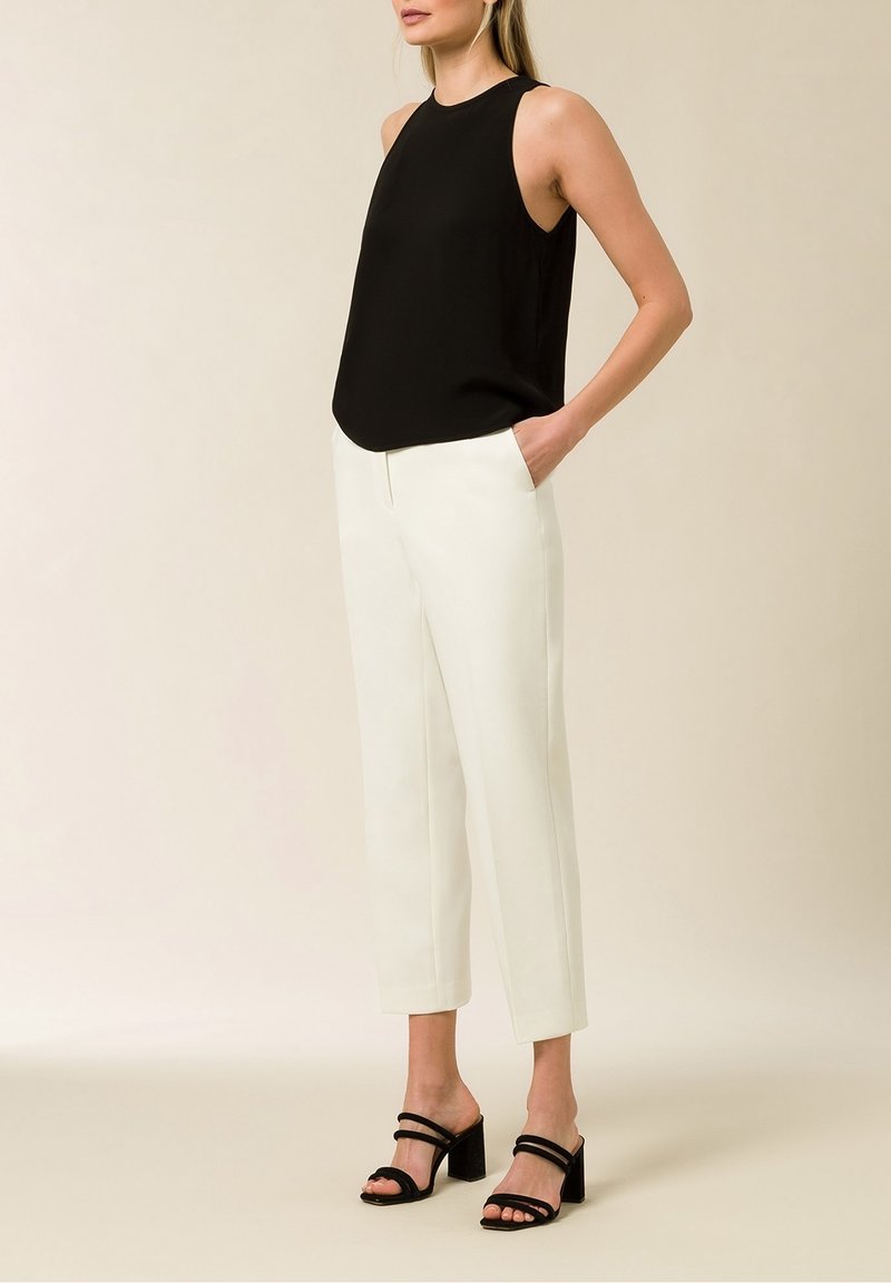 Femme portant un haut noir sans manches, un pantalon blanc court et des sandales à talons noires, debout avec une main dans la poche.