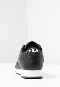 Black athletic shoe with a smooth synthetic upper, textured heel, white sole, and embroidered Fila logo in red and white at the back.