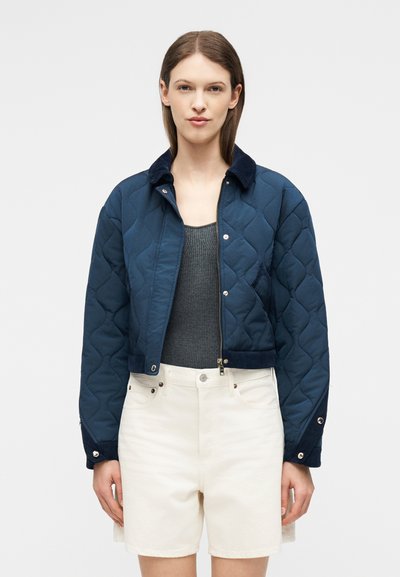 Femme portant une veste courte matelassée bleu marine par-dessus un débardeur côtelé gris foncé, associée à un short blanc taille haute, sur un fond uni.