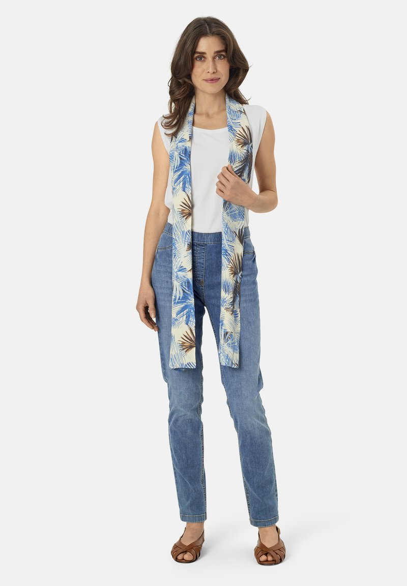 Blue and cream patterned scarf featuring palm leaves, worn over a white top and light blue jeans. The scarf is long and lightweight.