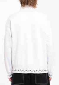 Sweat-shirt gris clair avec un col haut, des poignets côtelés et un motif noir brodé au niveau de l'ourlet. Tissu doux, coupe décontractée, design minimal.