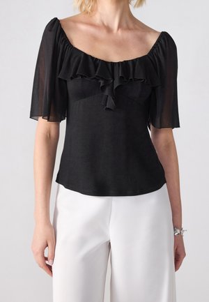 Zwarte off-shoulder blouse met ruches en doorschijnende mouwen tot op de elleboog, gedragen met witte, wijde broekspijpen op een lichte achtergrond.