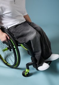 Črne široke kavbojke, bele čevlje in belo majico nosi oseba, ki sedi v invalidskem vozičku z zelenimi poudarki na modrim ozadju.