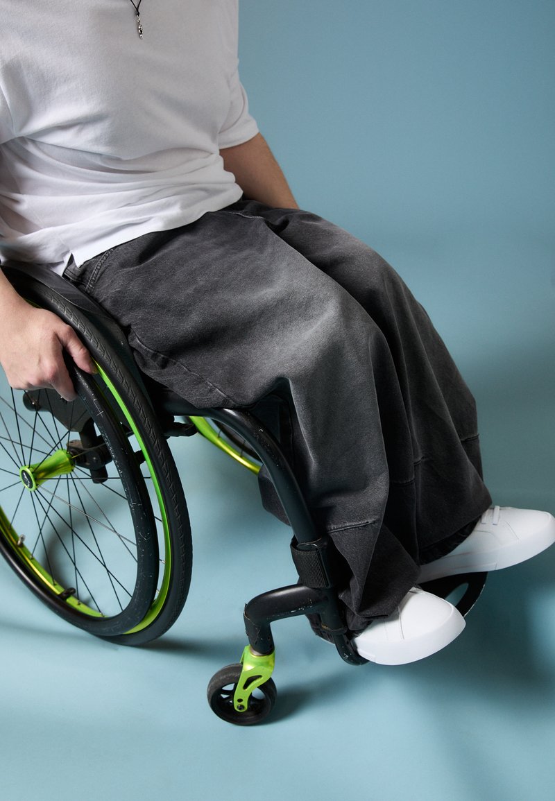 Črne široke kavbojke, bele čevlje in belo majico nosi oseba, ki sedi v invalidskem vozičku z zelenimi poudarki na modrim ozadju.
