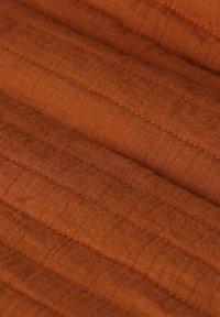 Tissu de couleur rouille avec une texture matelassée, présentant des lignes de couture parallèles uniformément espacées et une apparence douce, légèrement froissée.