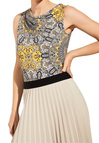 Ærmeløs top med et paisley-mønster i gult, sort og blåt; har en sort taljebånd og er parret med et plisseret beige skørt.