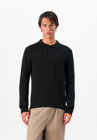 Polo preto de manga longa feito de tecido canelado, com uma pala de três botões e bainha reta. Usado com calças de tom bege claro.