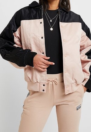 Femme portant une veste bomber beige et noire, un t-shirt noir, des colliers superposés, et un pantalon de survêtement beige avec une taille à cordon.