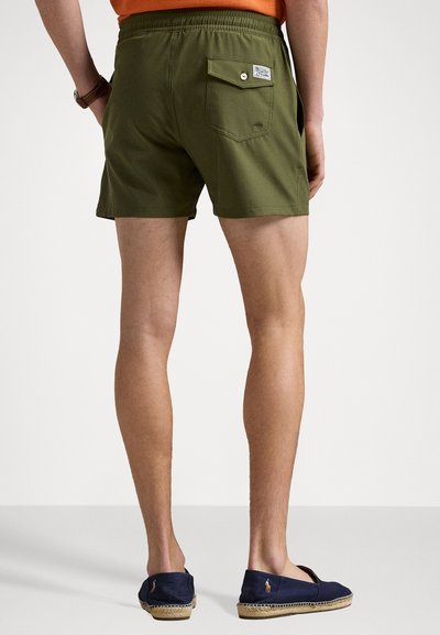 Calções verdes feitos de um tecido leve, com um bolso traseiro com fecho de botão e um pequeno rótulo da marca. Usados com alpargatas azul-marinho.