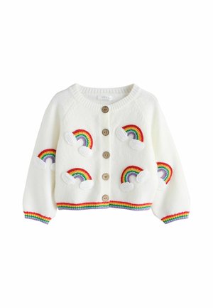 Peuter staat op een blauw tapijt, draagt een wit vestje met regenboog patches, brede blauwe spijkerbroek, witte sneakers, kijkt omhoog en opzij.