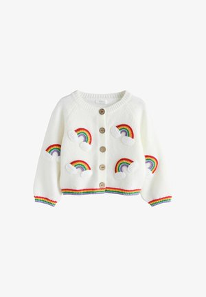 Kleinkind steht auf blauem Teppich, trägt weißen Cardigan mit Regenbogen-Patches, weite blaue Jeanshosen, weiße Turnschuhe, schaut nach oben und zur Seite.