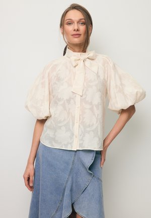 Vrouw die een doorschijnende blouse met bloemenprint, pofmouwen en een strikceintuur draagt, gecombineerd met een asymmetrische blauwe spijkerrok.